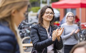 Frau mit Brille und schwarzer Jacke klatscht lächelnd. Im Hintergrund weitere Personen und ein roter Pavillon.