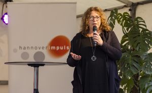 Frau mit lockigem Haar und Brille spricht in Mikrofon vor hannoverimpuls-Banner, trägt schwarzes Outfit mit Anhänger.