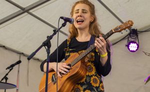 Junge Frau mit roten Haaren spielt akustische Gitarre und singt an einem Mikrofon auf einer Bühne.