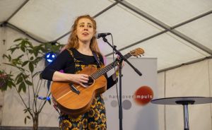 Junge Frau mit rotem Haar spielt Akustikgitarre und singt an einem Mikrofon in einem Zelt.