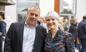Zwei Personen stehen lächelnd vor einem Gebäude, im Hintergrund eine Menschengruppe.