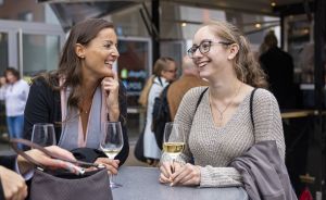 Zwei Frauen sitzen lachend an einem Tisch im Freien und halten je ein Glas Weißwein in der Hand.