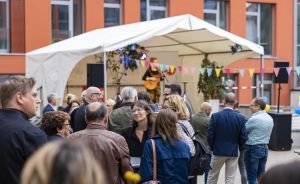 Menschengruppe auf einem Außenfest vor einem weißen Zelt mit Bühne, bunten Wimpeln und einem Musiker mit Gitarre.