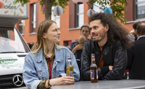 Zwei Personen sitzen an einem Tisch im Freien, halten Flaschen und unterhalten sich. Im Hintergrund ein weißer Van.