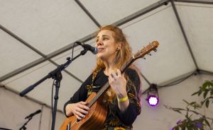 Frau mit langen Haaren spielt Akustikgitarre und singt in ein Mikrofon auf einer Bühne unter einem Zelt.