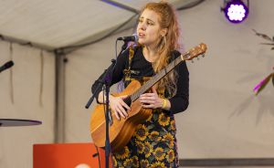 Junge Frau mit langen roten Haaren spielt akustische Gitarre und singt in ein Mikrofon auf einer Bühne.