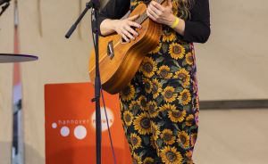 Junge Frau mit roten Haaren spielt Ukulele und singt auf einer Bühne. Sie trägt einen Sonnenblumen-Overall.