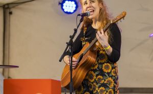 Frau mit rotem Haar spielt Akustikgitarre und singt in ein Mikrofon. Sie trägt ein Sonnenblumen-Overall auf einer Bühne.