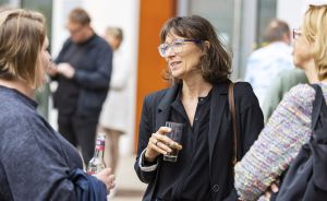 Frau mit Brille und schwarzer Jacke hält ein Glas und spricht mit zwei Personen bei einer Veranstaltung im Freien.