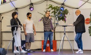 Vier Personen auf einer Bühne unter einem Zelt, einer spricht in ein Mikrofon, im Hintergrund Scheinwerfer und eine Pflanze.