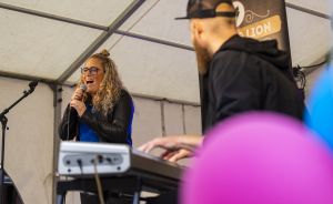 Frau mit lockigem Haar singt ins Mikrofon auf einer Bühne, Mann spielt Keyboard, bunte Luftballons im Vordergrund.