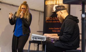 Frau mit blonden Locken singt ins Mikrofon, Mann mit Kappe spielt Keyboard. Banner „Curly & Li… Duo-Live" im Hintergrund.