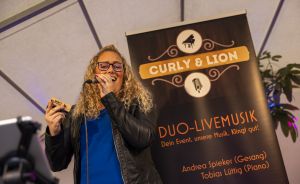 Frau mit Locken und Brille singt ins Mikrofon vor einem Banner „Curly & Lion Duo-Livemusik" mit schwarzer Lederjacke.