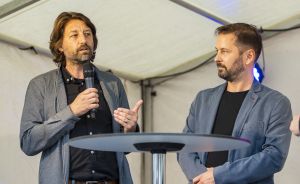 Zwei Männer in grauen Blazern stehen an einem Stehtisch; einer spricht in ein Mikrofon.