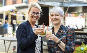 Zwei Frauen stoßen lächelnd mit Cocktailgläsern an. Im Hintergrund ein Foodtruck und weitere Personen.