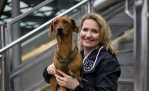 Blonde Frau sitzt auf Treppe und hält braunen Dackel mit grünem Geschirr im Arm. Beide schauen in die Kamera.