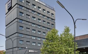 Modernes dunkles Hochhaus mit Aufschrift „NYCE Hotel", Straßenlaterne, Baum und Zebrastreifen im Vordergrund.
