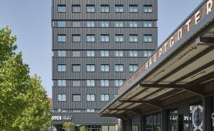 Modernes dunkles Hochhaus hinter überdachtem Bahnsteig "Hauptguter", Bäume, Graffiti, Fußweg mit Streifen.