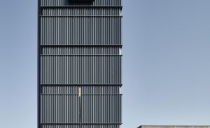Modernes dunkles Hochhaus mit vertikaler Metallverkleidung und "AYCE Hotel"-Schild auf dem Dach, blauer Himmel.