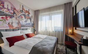 Hotelzimmer mit Doppelbett, roten Kissen, großem Wandgemälde, Schreibtisch, TV und Fenster mit Vorhängen.