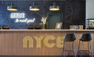 Hotelrezeption mit Holzverkleidung, „NYCE"-Logo, Neonschrift, zwei schwarzen Barhockern und Stadtkartenillustration.