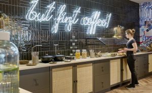 Frau steht an einer Küchentheke mit dunklen Fliesen. Neon-Schriftzug „but first coffee!" an der Wand.