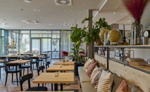 Helles Restaurant mit Holztischen, schwarzen Stühlen, Sofa mit Kissen, Zimmerpflanzen und Glasfront zum Außenbereich.