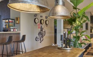 Industrielle Hängelampen aus Metall über langem Holztisch, Barhocker, Wanduhren, Grünpflanzen in modernem Café-Interieur.