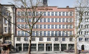 Mehrstöckiges Stadtgebäude mit rosa Fassade, Aufschrift „Kurierhaus" und „Motel One", flankiert von kahlen Bäumen.
