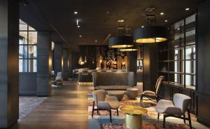Hotellobby mit dunklen Wänden, goldenen Rundschemeln, grauen Sesseln, großen Hängelampen und gemustertem Teppich.