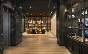 Moderner Hotelflur mit Holzboden, schwarzer Marmorwand, Aufzug, Bücherregal und goldenen Sesseln im Hintergrund.