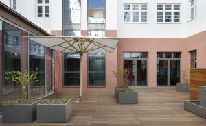 Holzterrasse mit großem Sonnenschirm, grauen Pflanzkübeln und Glastüren an einem rosa-weißen Gebäude.