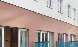 Mehrgeschössiges Gebäude mit rosa Sockel, weißer Fassade, blauen Glasbrüstungen und türkisfarbenen Liegestühlen auf Terrassen.