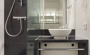 Modernes Badezimmer mit schwarzen Fliesen, Glasdusche mit Regendusche und weißem Aufsatzwaschbecken auf dunkler Ablage.