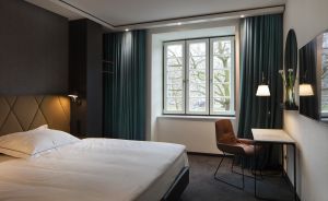 Hotelzimmer mit Doppelbett, grünen Vorhängen, Fenster, Schreibtisch mit Stuhl und Wandspiegel.