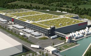 Luftbild eines großen Lagergebäudes mit begrüntem Dach, Laderampen, geparkten LKWs und angrenzendem Kanal.