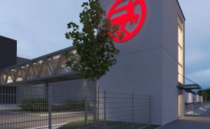Modernes Industriegebäude bei Dämmerung mit rotem Firmenlogo auf Turm, Metallzaun und Brückenverbindung.