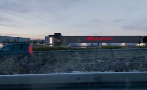 Rossmann-Gebäude bei Dämmerung mit beleuchtetem Schriftzug, davor eine Straße mit einem fahrenden Auto.