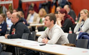 Mehrere Personen sitzen an langen Tischen in einem Saal und hören aufmerksam zu. Auf den Tischen liegen Papiere und Flaschen.