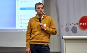 Mann in gelbem Pullover spricht mit Mikrofon vor Präsentationsfolie und "hannoverimpuls"-Banner.