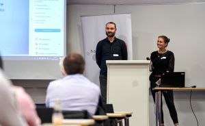Zwei Personen stehen vor einer Projektionsleinwand mit Adobe-PDF-Menü und einem Roll-up-Banner in einem Seminarraum.