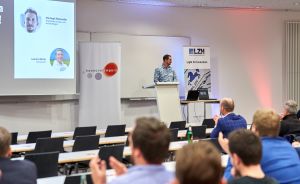 Mann am Rednerpult hält Vortrag vor Publikum in Konferenzraum; Leinwand mit Portraits, Banner von hannoverimpuls und LZH.