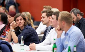 Mehrere Personen sitzen an Tischen in einem Veranstaltungsraum und hören aufmerksam zu. Flaschen stehen auf den Tischen.