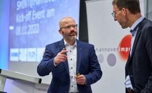 Zwei Männer in Anzügen bei einer Präsentation. Der linke trägt ein Headset-Mikrofon und zeigt mit dem Finger.