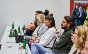 Mehrere Personen sitzen in einer Reihe an einem Tisch mit Glasflaschen und hören aufmerksam zu.