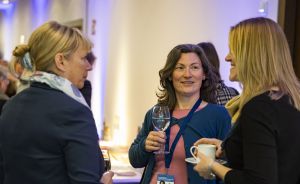Drei Frauen unterhalten sich bei einer Veranstaltung; eine hält ein Weinglas, eine andere eine Tasse. Eine trägt ein Namensschild.