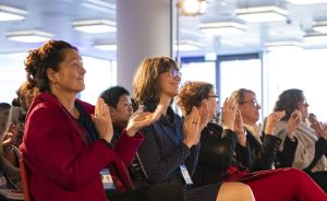 Mehrere Frauen sitzen in einer Reihe und klatschen lächelnd in einem hell beleuchteten Konferenzraum.