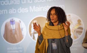 Frau mit lockigem Haar und gelbem Schal gestikuliert vor einer deutschen Präsentationsfolie mit Modebildern.