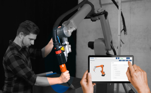 Mann in kariertem Hemd bedient Roboterarm; Hände halten Tablet mit 3D-Robotermodell auf dem Bildschirm.