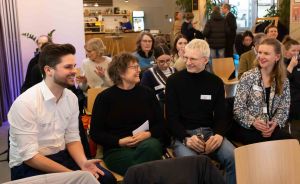 Vier Personen sitzen in Stuhlreihen und unterhalten sich lachend. Im Hintergrund stehen weitere Menschen in einem Raum.
